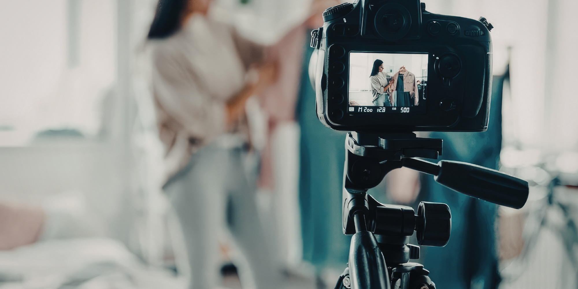 content-creator-social-video-riprese Vista ravvicinata attraverso lo schermo di una videocamera DSLR professionale montata su cavalletto, che inquadra una giovane content creator donna vestita in denim mentre si muove in un ambiente lifestyle luminoso e sfocato.